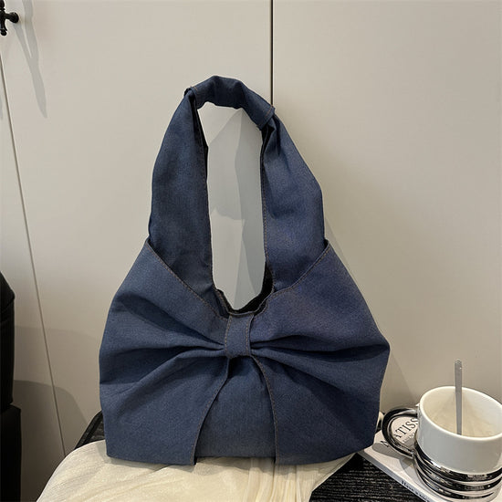 Dark Blue denim bow shoulder bag with oversized bow resting on a table beside a coffee mug
