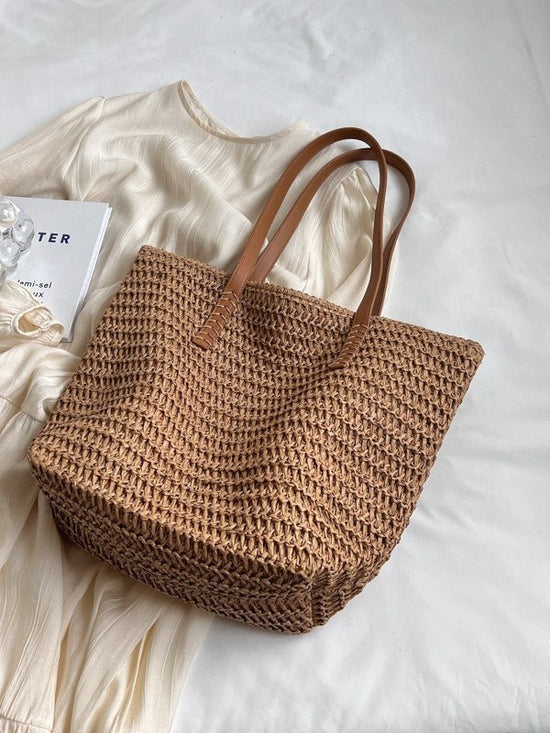 Boho Baecation Khaki woven straw tote bag with two brown handles, flat lay on white bedding beside a cream dress
