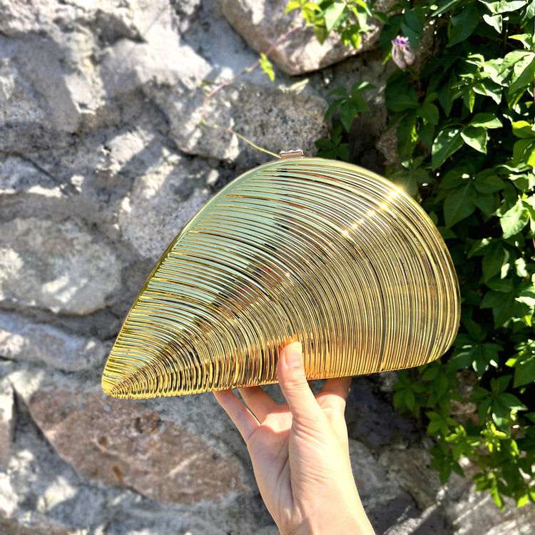 Hand holding a gold ribbed fan-shaped acrylic clutch against a rustic stone wall with green foliage
