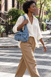 Woman in a white shirt and tan pants crossing a city street with a Blue denim bow shoulder bag on her shoulder