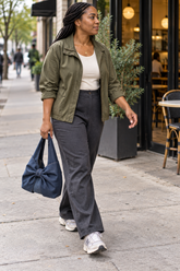 Woman in an olive jacket and gray pants walking on a sidewalk carrying a Dark Blue denim bow shoulder bag