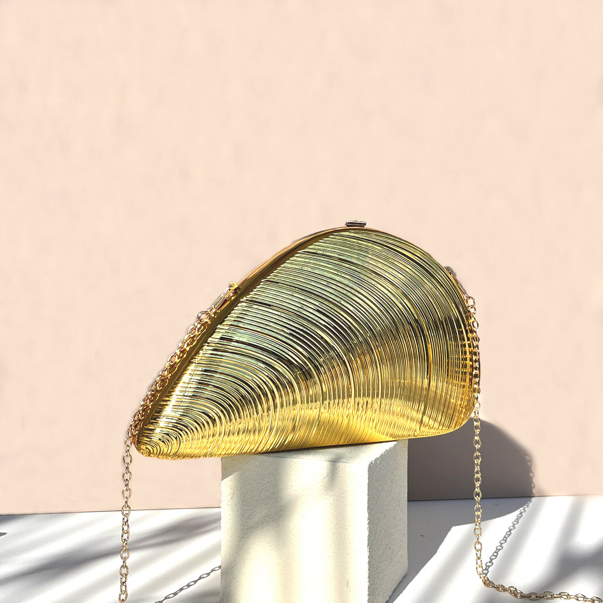 Gold ribbed fan-shaped acrylic clutch with gold chain strap displayed on a white pedestal against a pale beige background